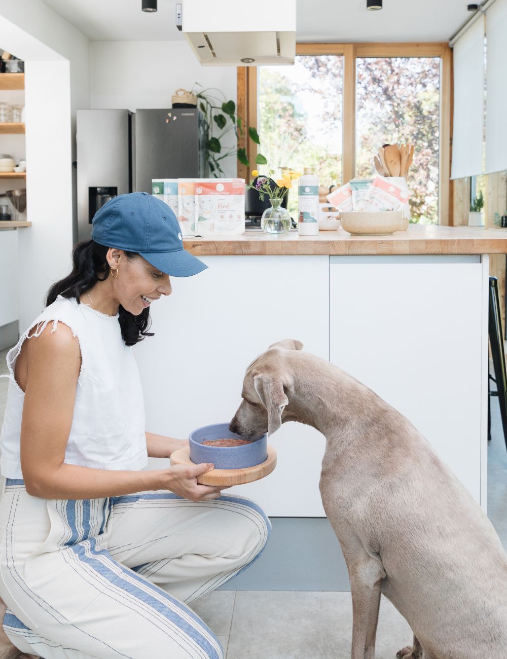Lächelnde Frau, die kniend ihrem Hund Futter anbietet. Dahinter eine weiße moderne Küche mit großem Fenster
