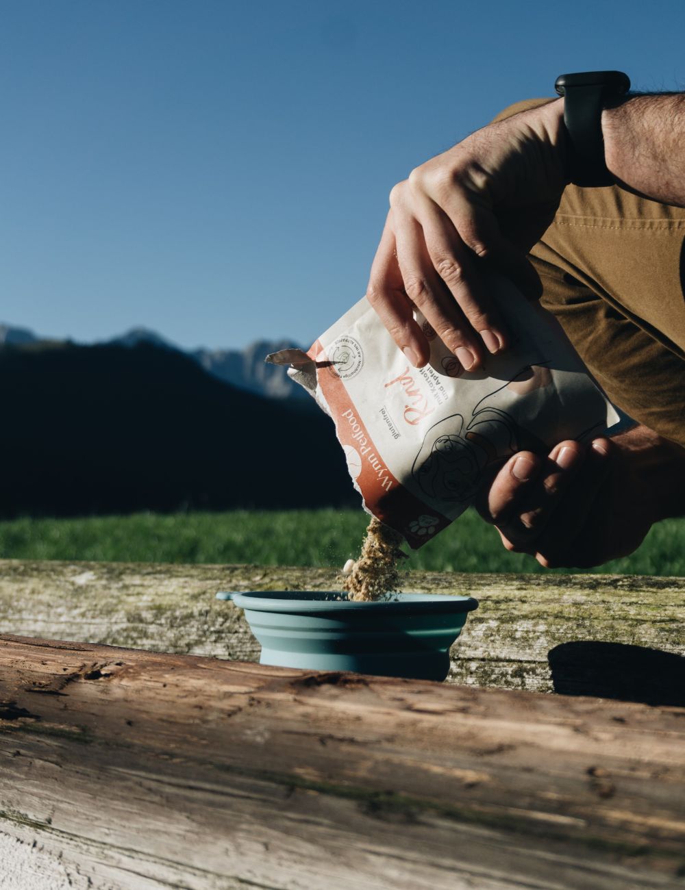 Futter das in einen Napf geleert wird. Im Hintergrund sind Berge zu erkennen und ein blauer Himmel.