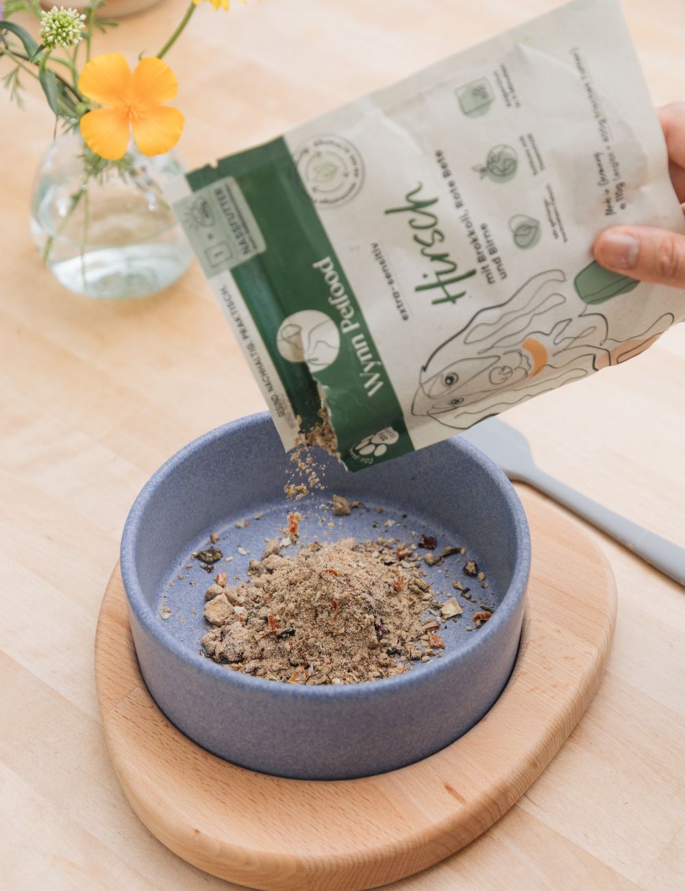 Futtertüte, die gerade in Napf ausgekippt wird. Der Napf hat einen Untersetzer aus Holz.